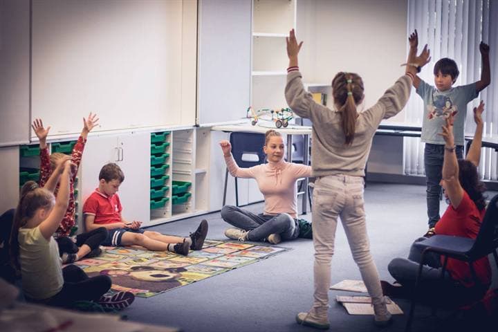 Children raising their hands during a performance-style classroom activity.