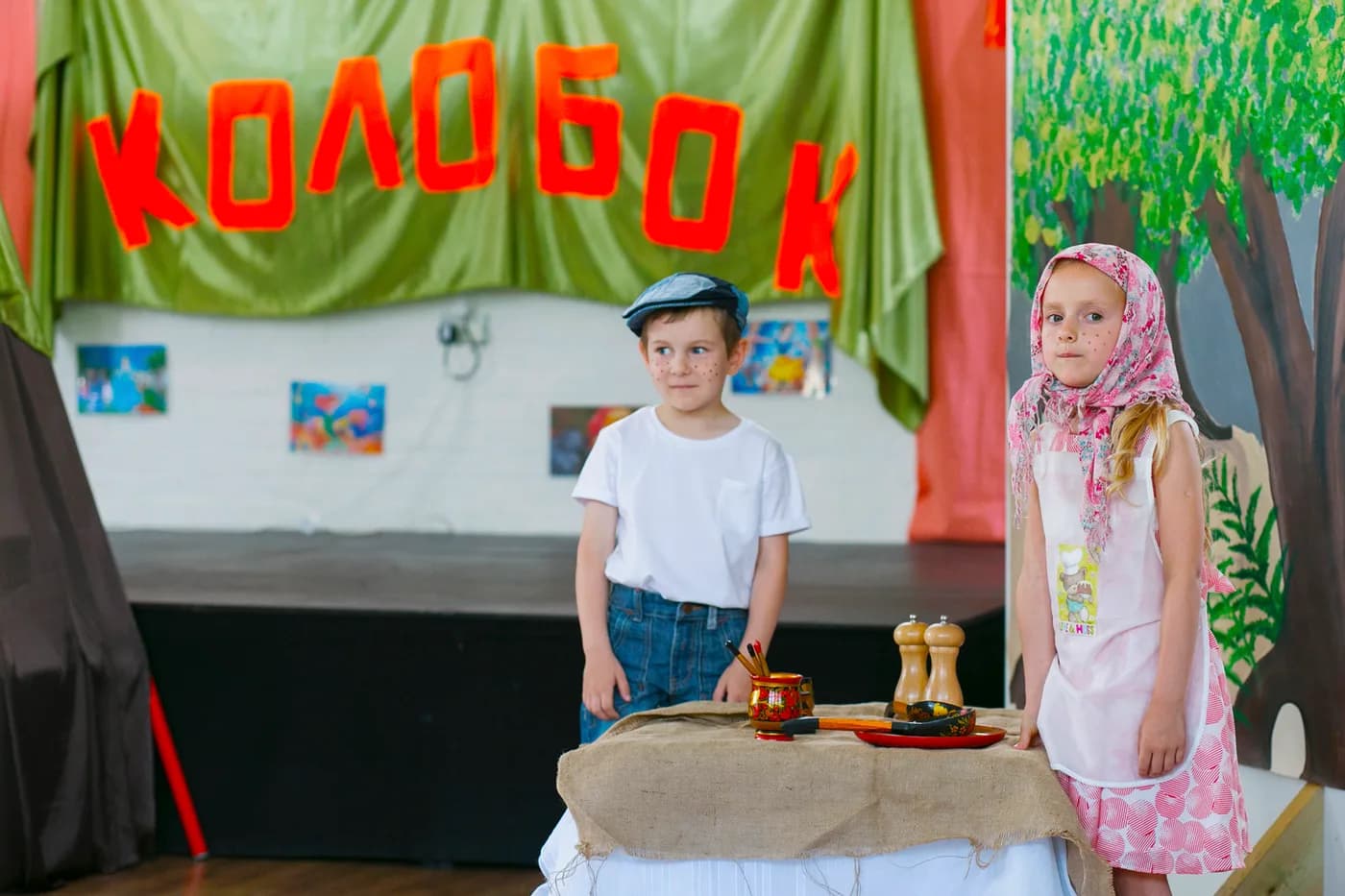 Children performing a Kolobok stage scene with props.